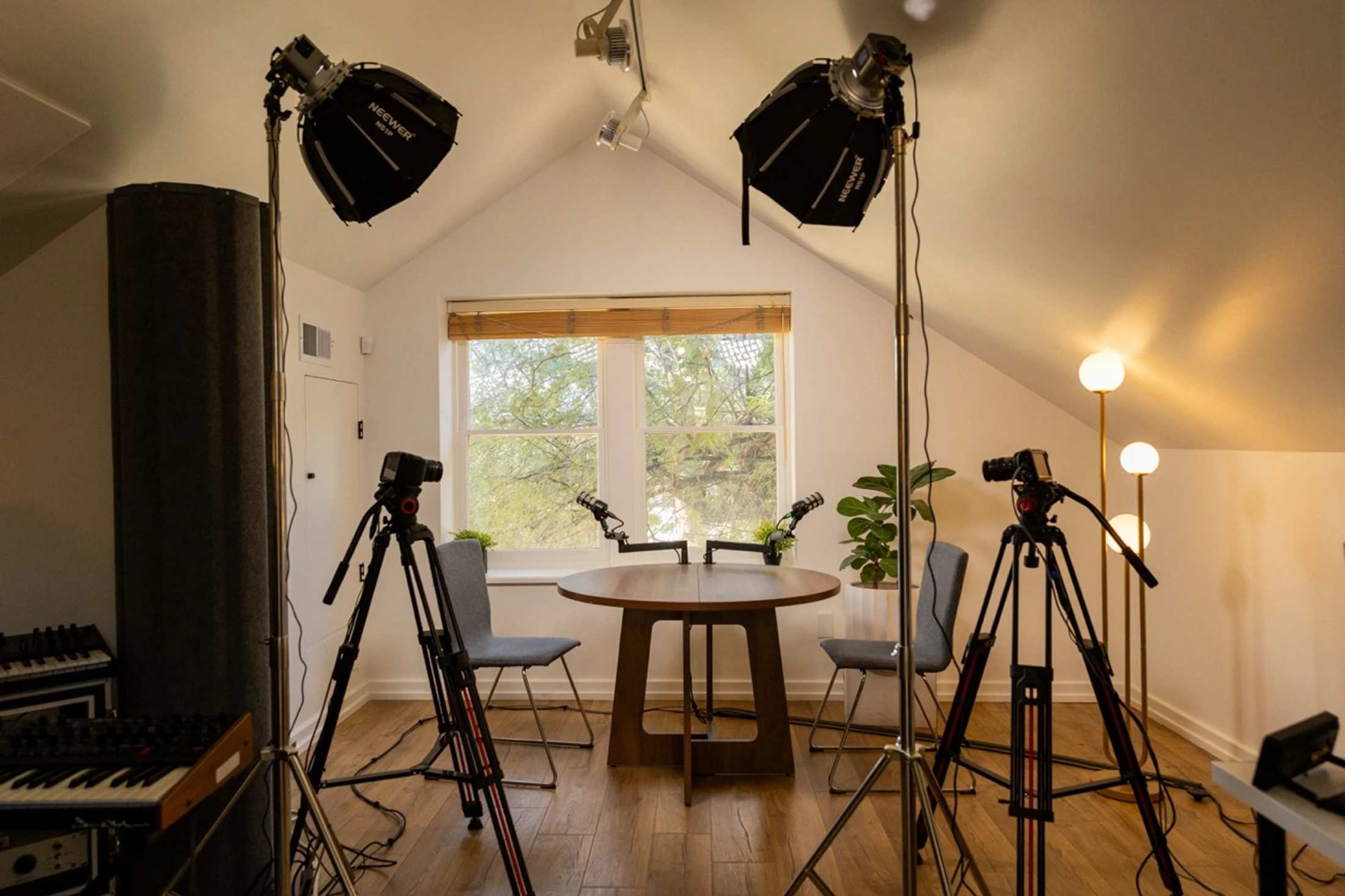 A professional recording studio with a table, two chairs, and two microphones for a podcast. The room is equipped with cameras on tripods and softbox lights.
